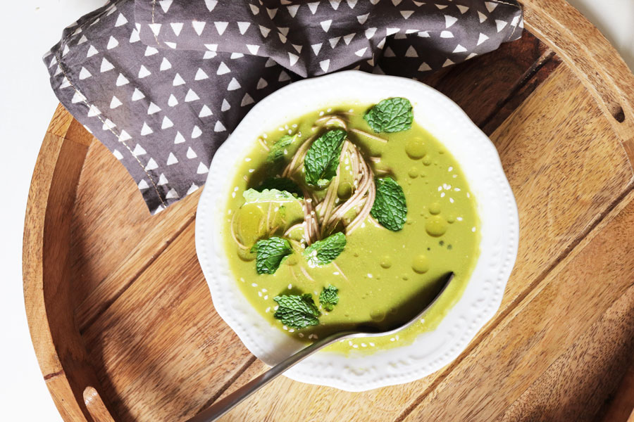 A wooden tray with a bowl of green pesto soup, topped with soba noodles and mint leaves